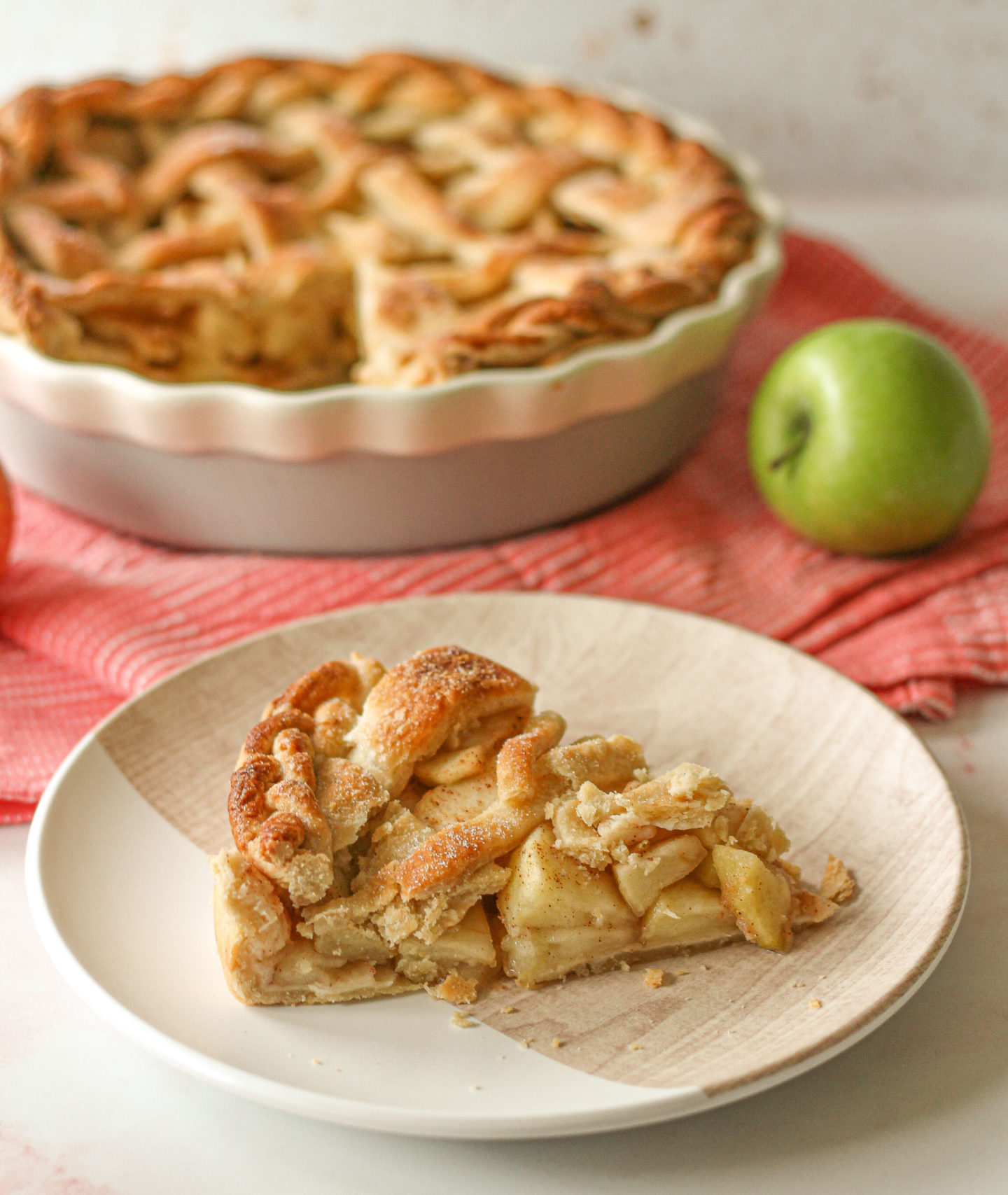 Slice of lattice apple pie with whole lattice apple pie blurred in background