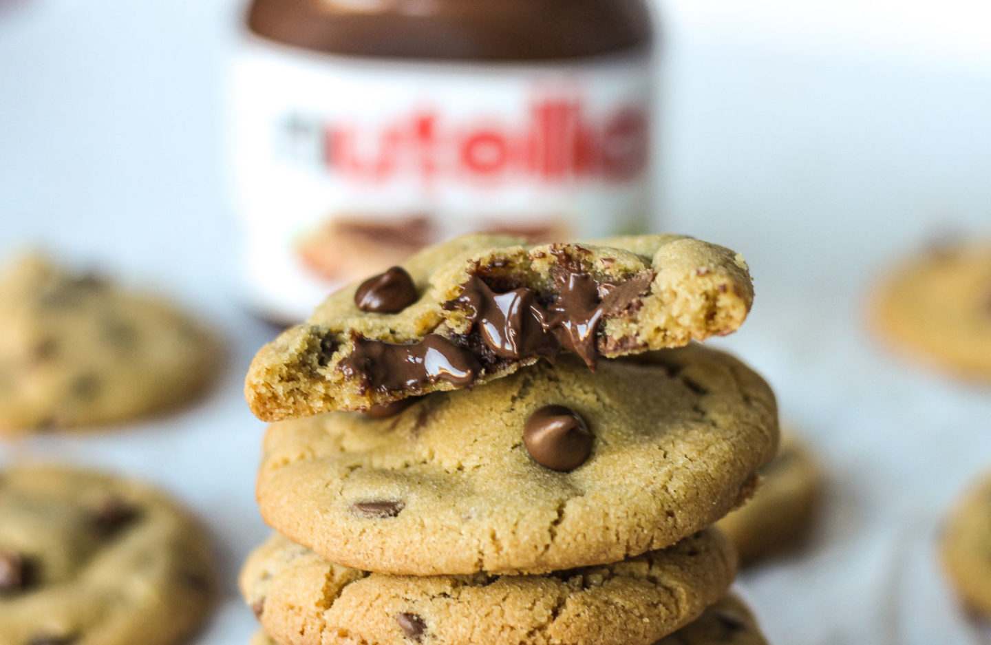 half a Nutella stuffed chocolate chip cookie showing melted Nutella inside, sat on top of tower of cookies with jar of Nutella in background