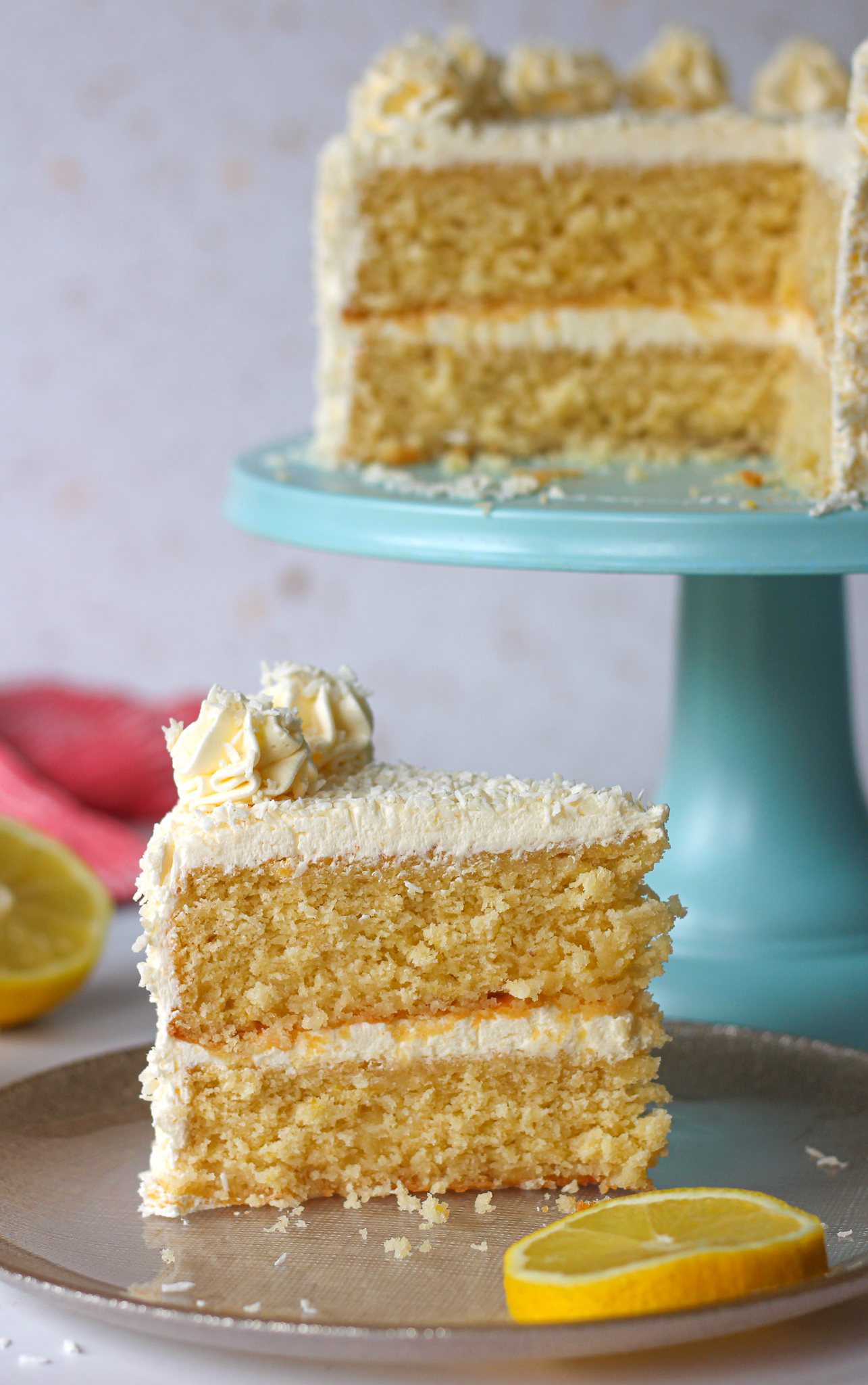 close up of a slice of lemon coconut layer cake with rest of cake on cake stand blurred in background 