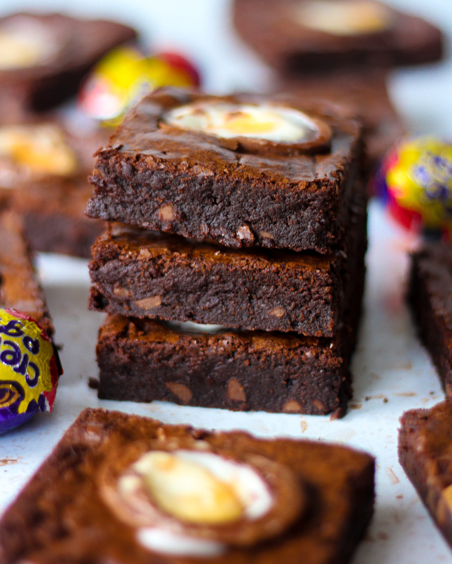 stack of three chewy creme egg brownies surrounded by other brownie slices and Creme Eggs blurred in background