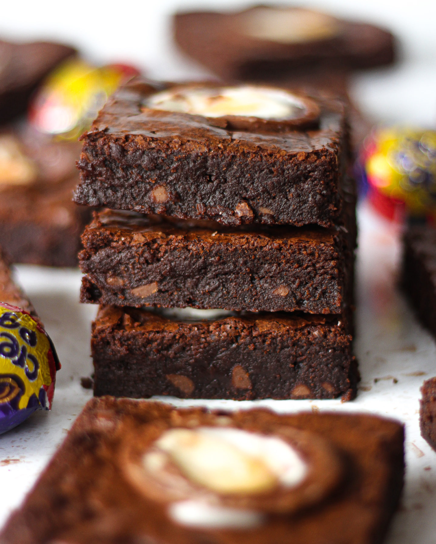 stack of three chewy creme egg brownies surrounded by other brownie slices and Creme Eggs blurred in background