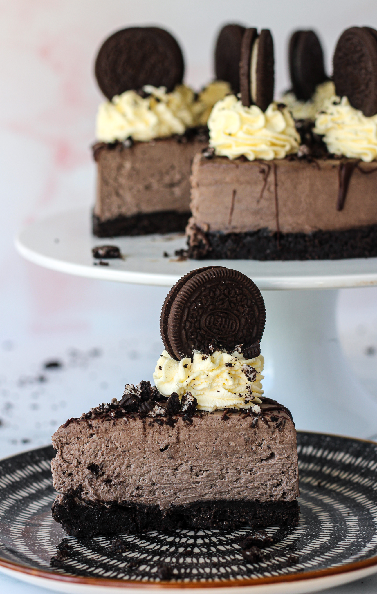 close up of a slice of no-bake Oreo cheesecake on a serving plate with rest of cheesecake on a cake stand in the background