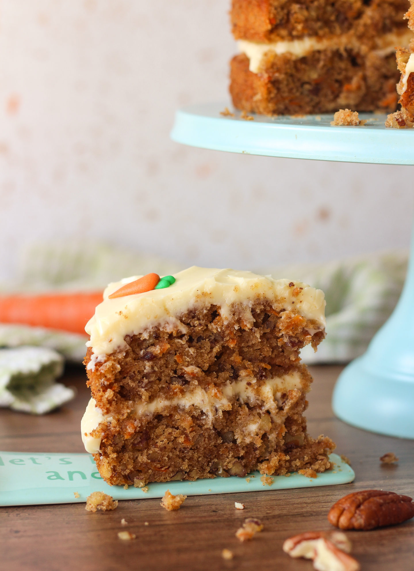 close up of a slice of carrot cake with the rest of the unsliced carrot cake on a blue stand in the background